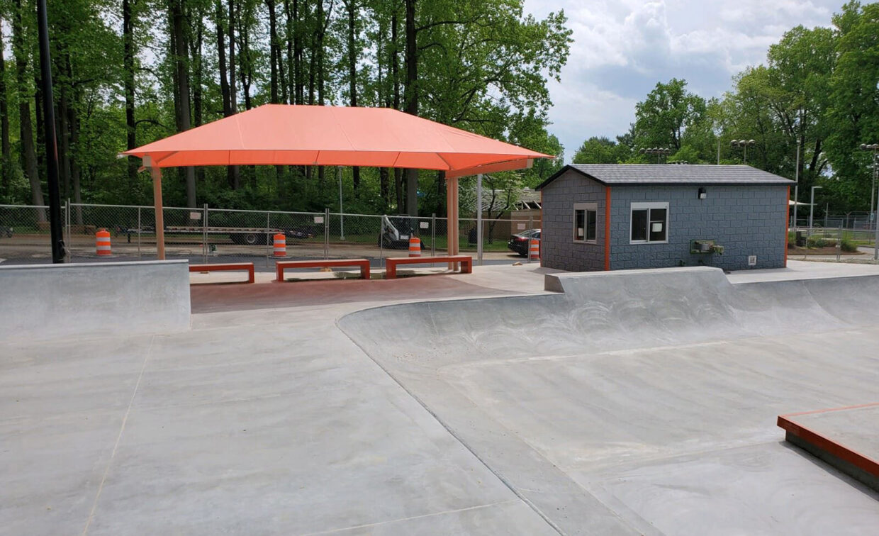 A concrete skatepark with ramps, an orange-roofed shade structure, benches, and a small gray building, surrounded by trees and a chain-link fence.