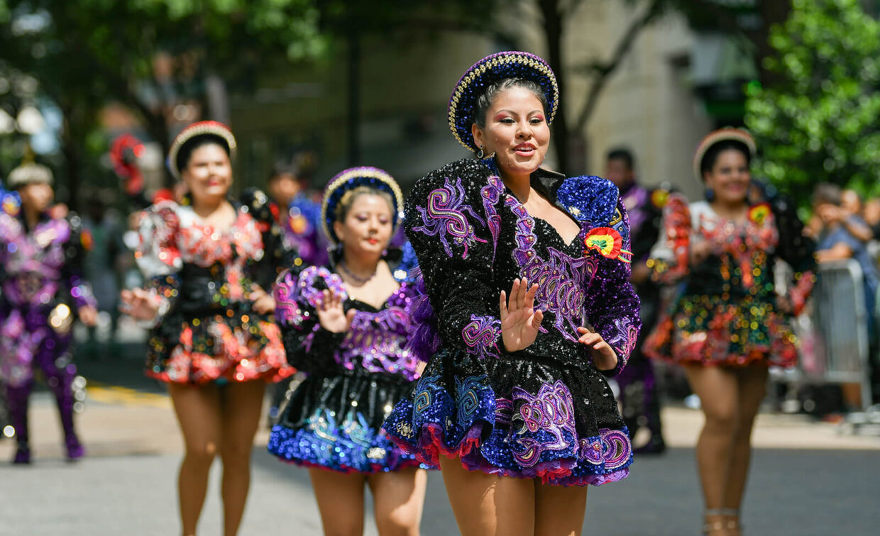 A group of women in colorful, sequined costumes dance in a street parade during daylight, with trees and spectators in the background.