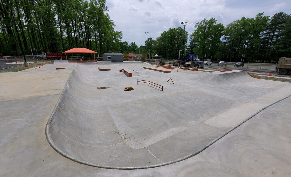 A concrete skatepark with ramps, rails, and ledges, surrounded by trees and some construction equipment in the background.