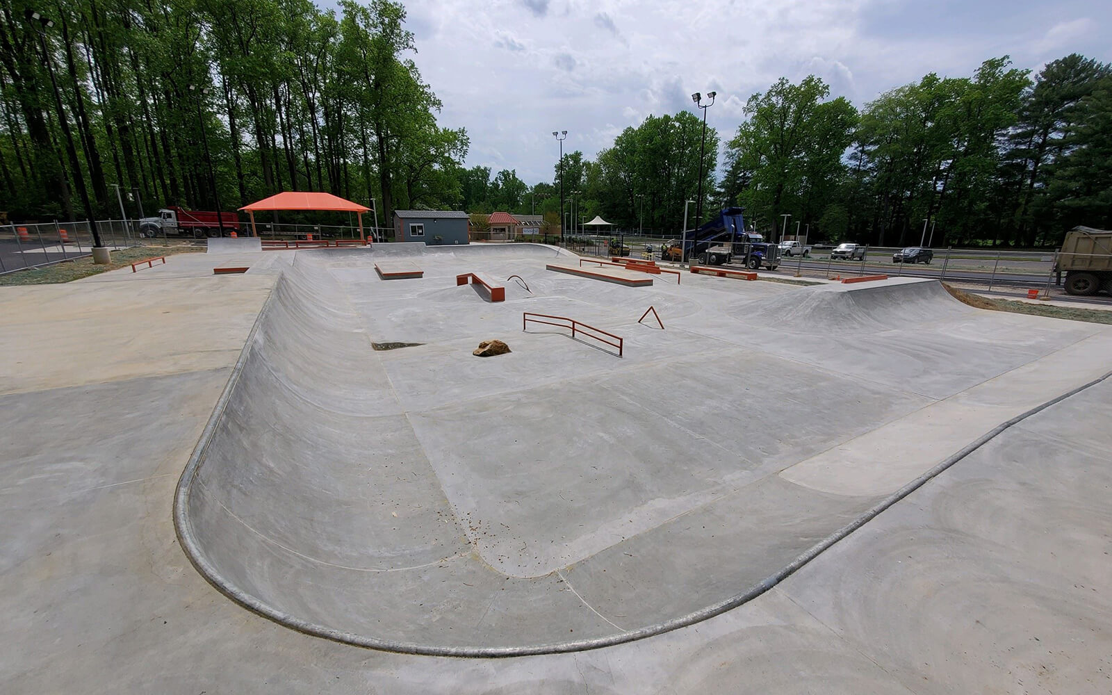 A concrete skatepark with ramps, rails, and ledges, surrounded by trees and some construction equipment in the background.