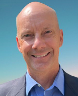 A middle-aged man with a bald head, wearing a blue collared shirt and dark jacket, smiling against a clear blue sky background.