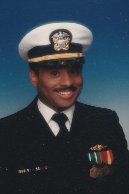 A man in a U.S. Navy dress uniform and officer’s hat, smiling, with medals and ribbons on his chest against a blue gradient background.