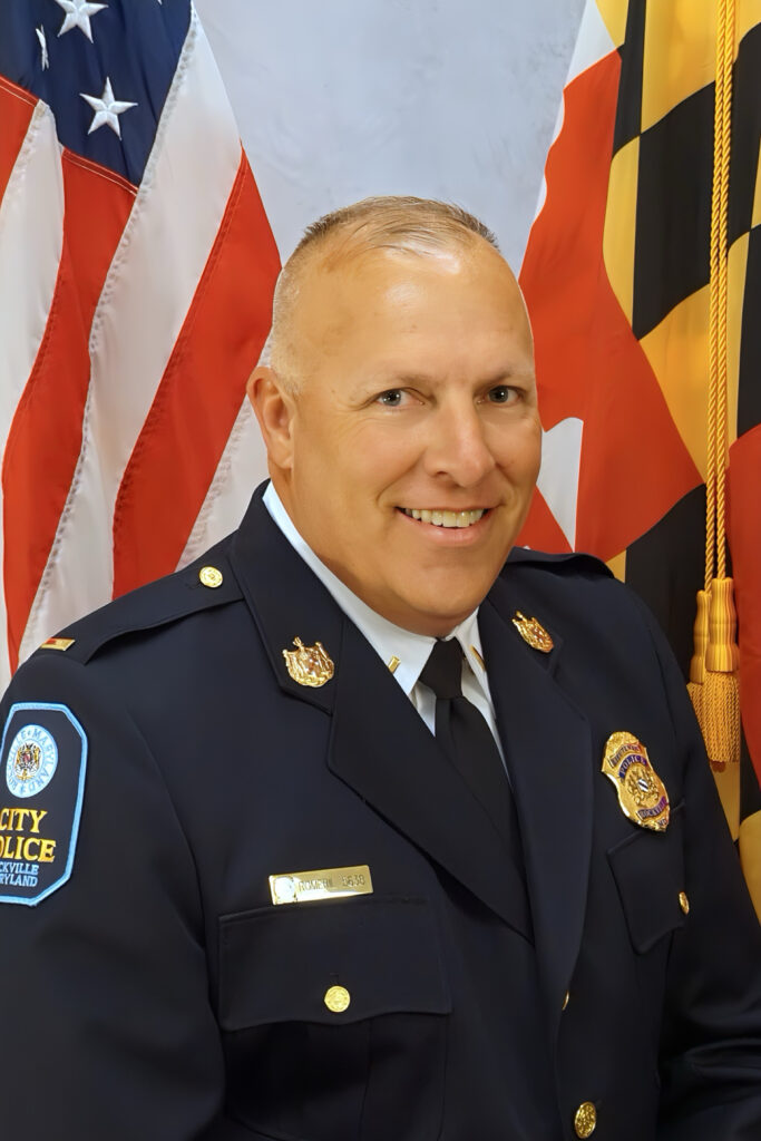A police officer in formal uniform poses in front of the American and Maryland state flags, smiling for an official portrait.