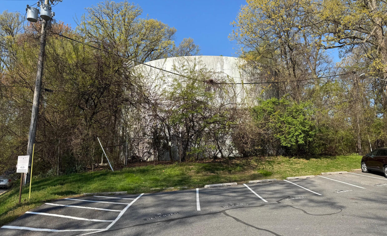 A large white cylindrical water tower is partially hidden by trees and bushes near a small parking lot with marked spaces on a sunny day.
