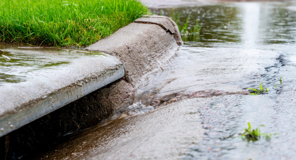 Rainwater flows from a grassy sidewalk curb into a street gutter during a heavy rainfall, causing runoff and minor flooding.
