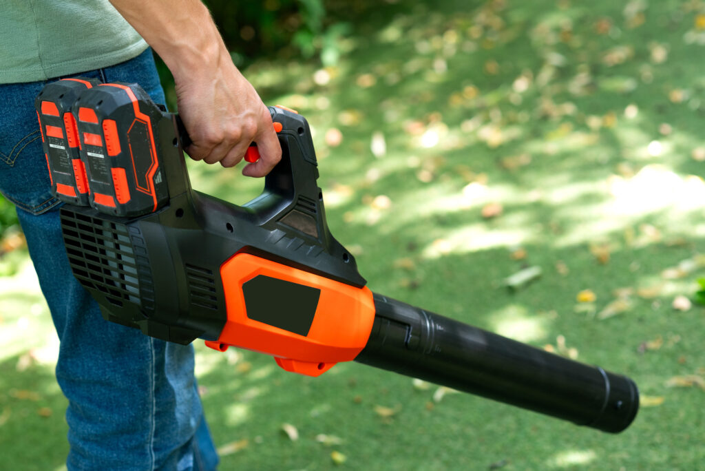 Person holding a battery-powered leaf blower outdoors, standing on green grass with scattered leaves in the background.