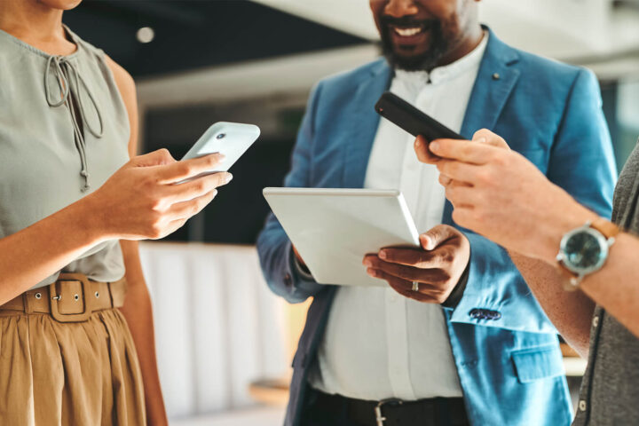 Three people stand close together, each holding a digital device—a smartphone or tablet—while interacting in a modern indoor setting.