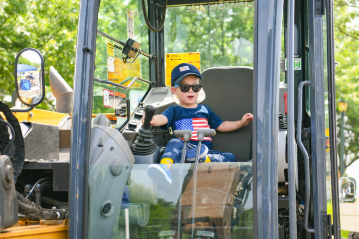 A young child wearing sunglasses and a hat sits in the driver’s seat of a construction vehicle, holding the controls.