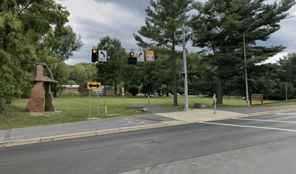 A street intersection with traffic lights, a yellow left turn sign, a sidewalk, grass, trees, and a large stone sculpture on the left.