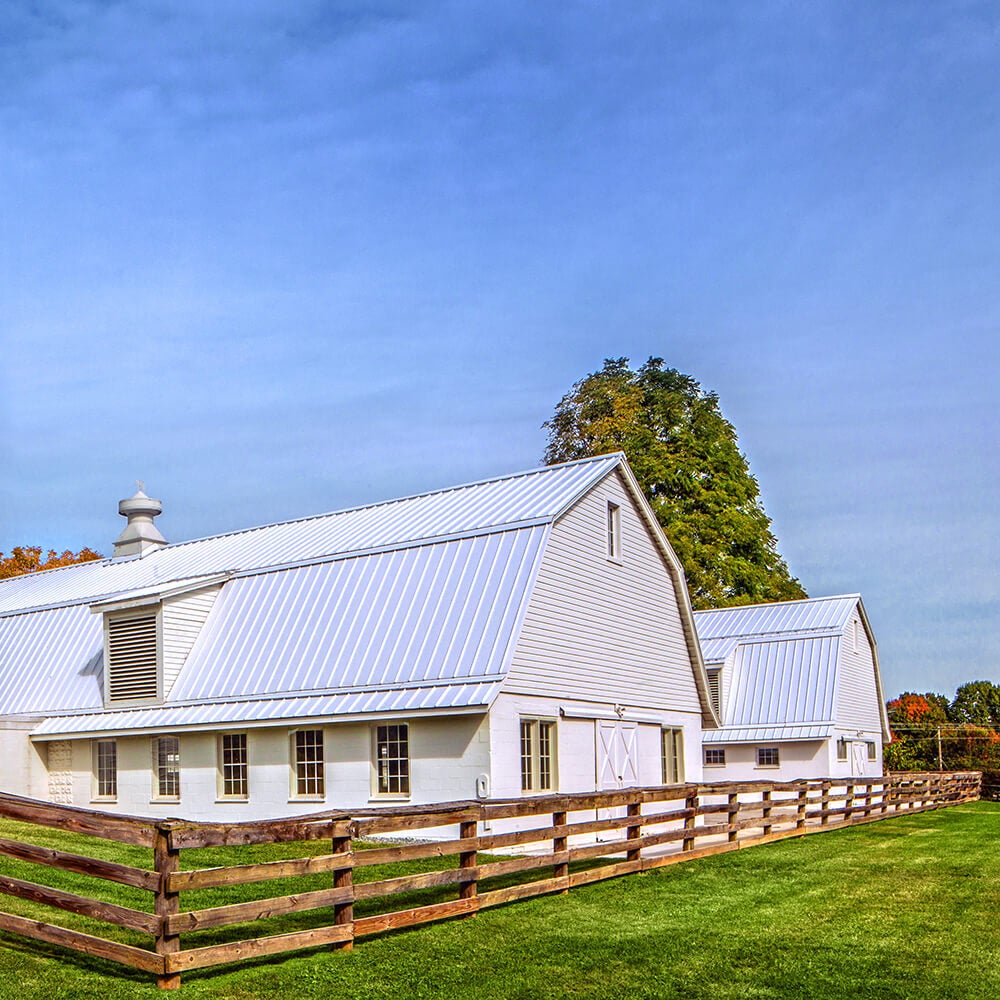 A white barn with two tall silos and a wooden fence sits on a grassy field under a blue sky.