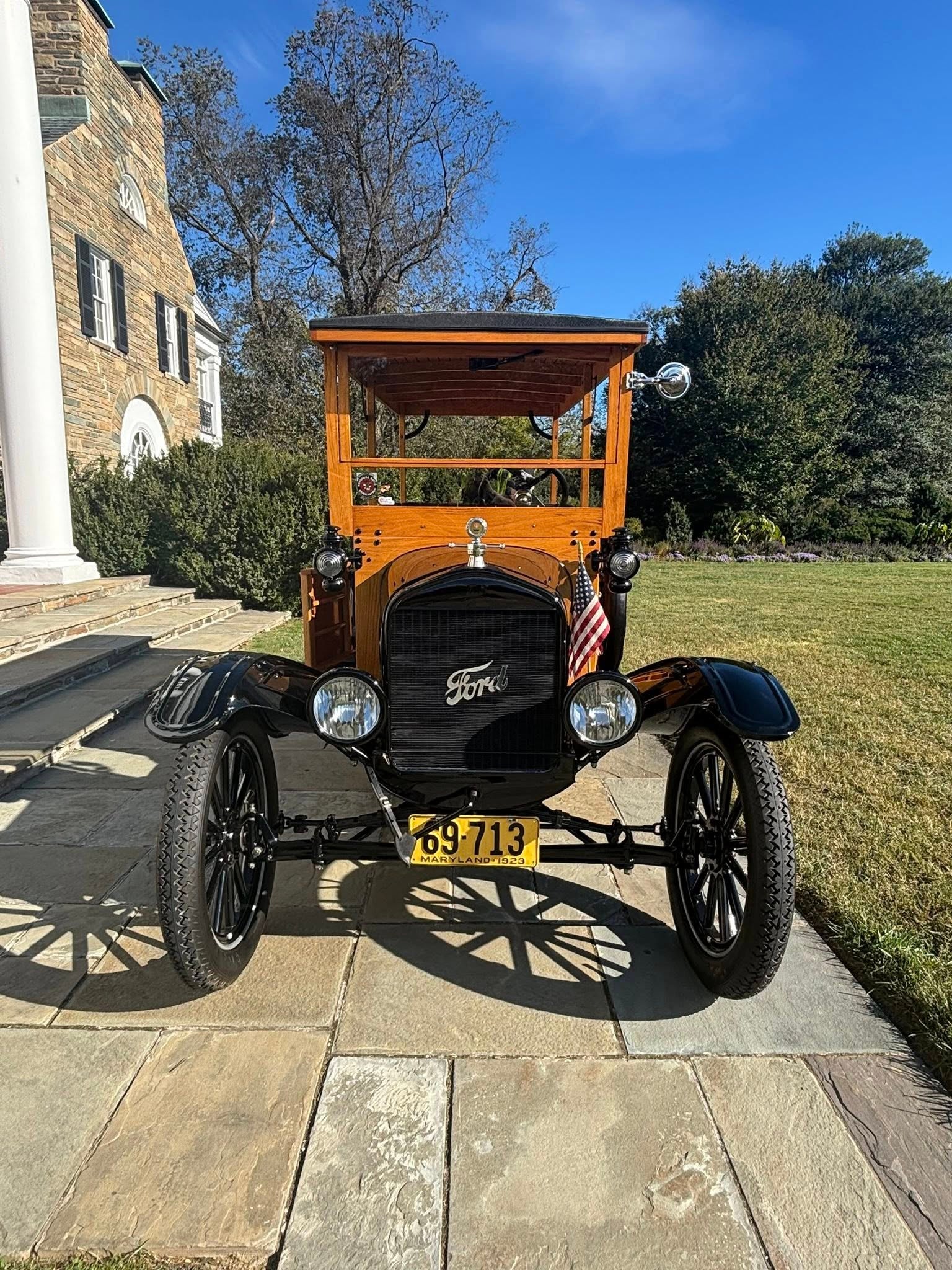A vintage black Ford Model T with wooden detailing is parked on a stone driveway in front of a house, displaying a yellow license plate and a small American flag.