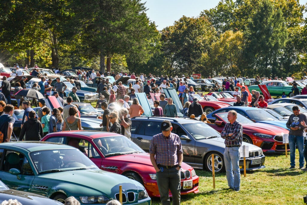 A large crowd gathers at an outdoor car show with many parked cars on display, some with open hoods, surrounded by trees on a sunny day.