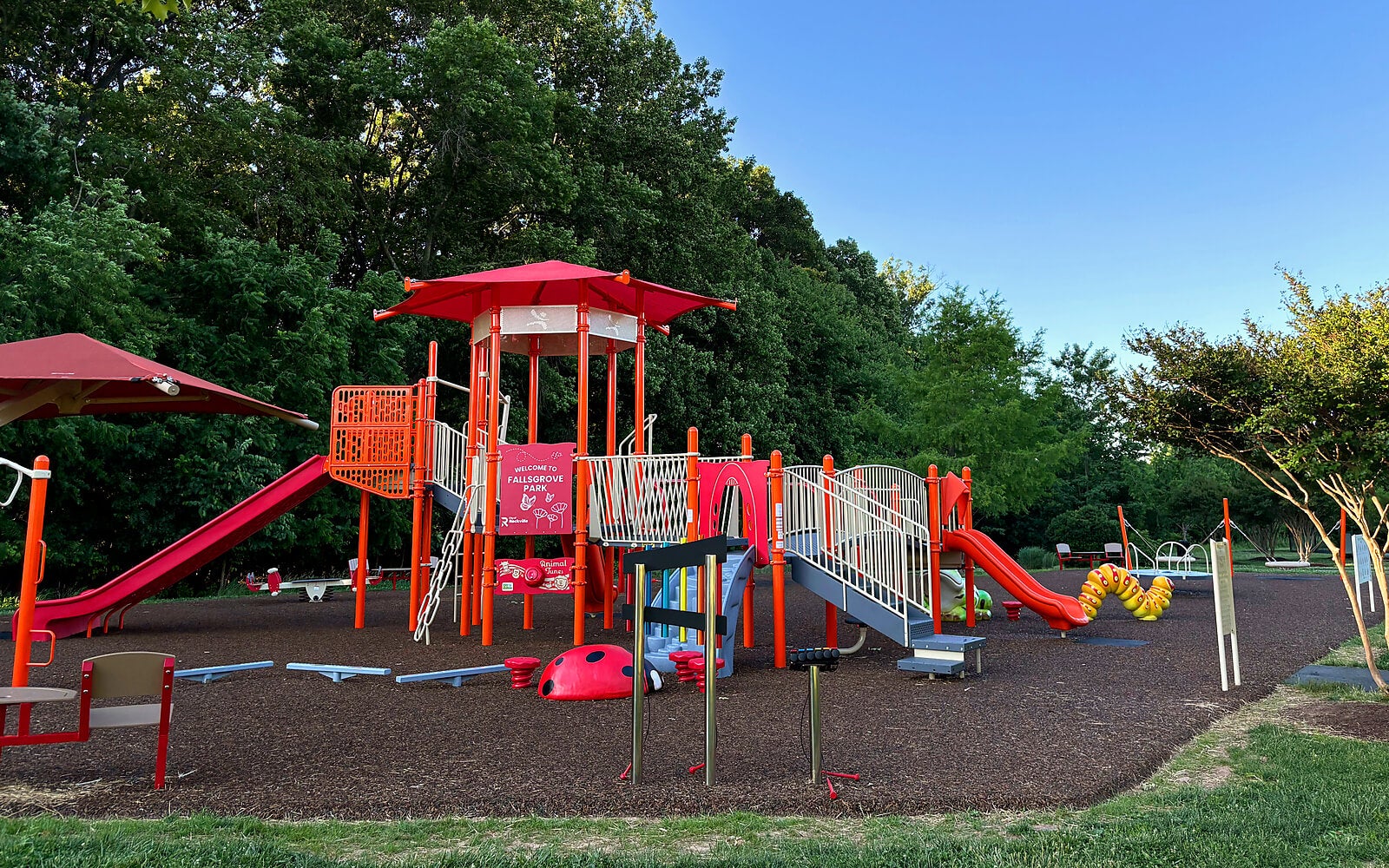 A playground with red and white climbing structures, slides, and a bridge, surrounded by trees and grass under a clear blue sky.