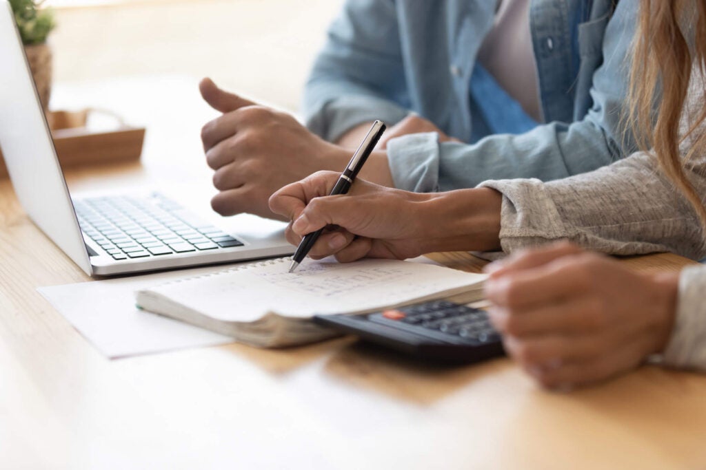 Two people work at a desk with a laptop, notebook, pen, and calculator; one writes while the other gives a thumbs up.
