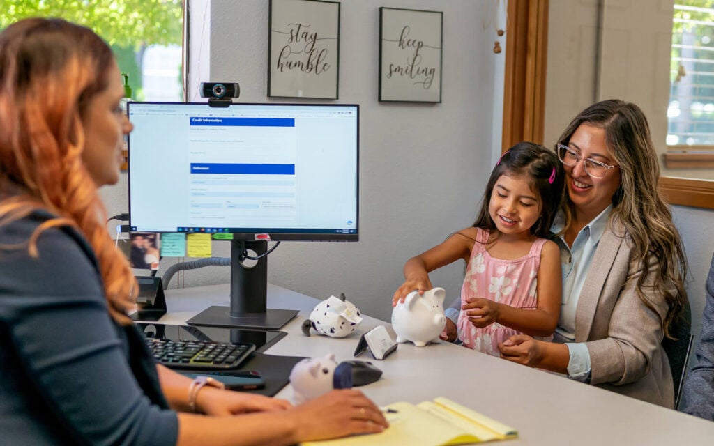 A woman and child interact with piggy banks at an office desk while another woman consults with them, with a computer monitor and paperwork visible.