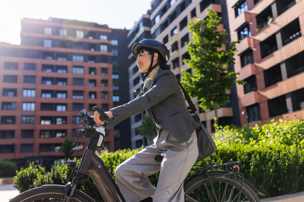 A person in business attire and a helmet rides a bicycle in an urban area with modern apartment buildings and greenery.