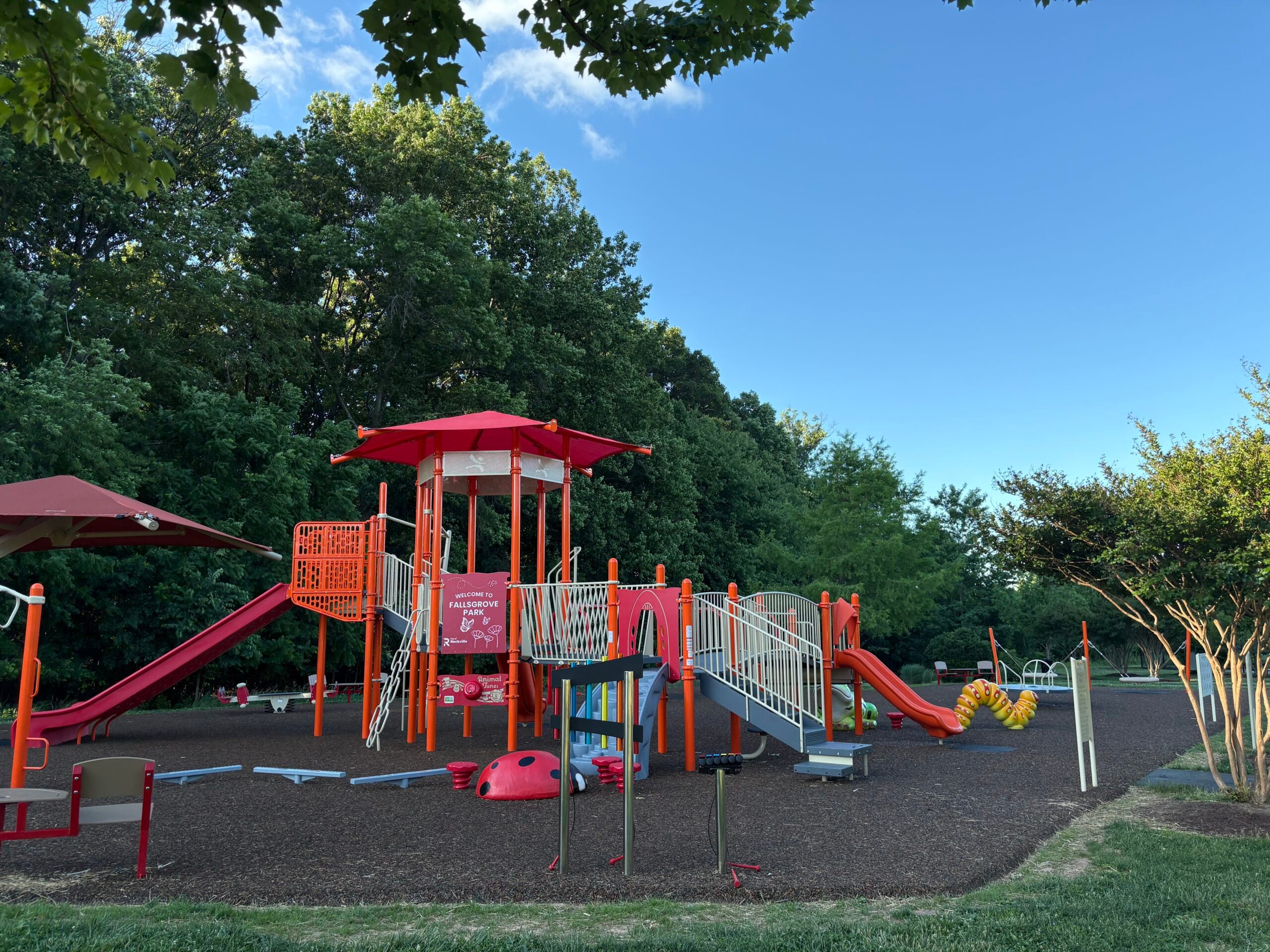 A playground with red and white climbing structures, slides, and a bridge, surrounded by trees and grass under a clear blue sky.