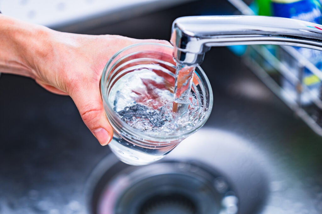 A hand holds a glass under a running kitchen faucet, filling it with water above a sink.