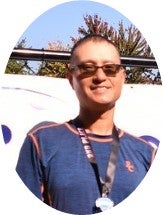 A person wearing sunglasses and a blue shirt with an event lanyard, standing outdoors in front of a white banner and trees.