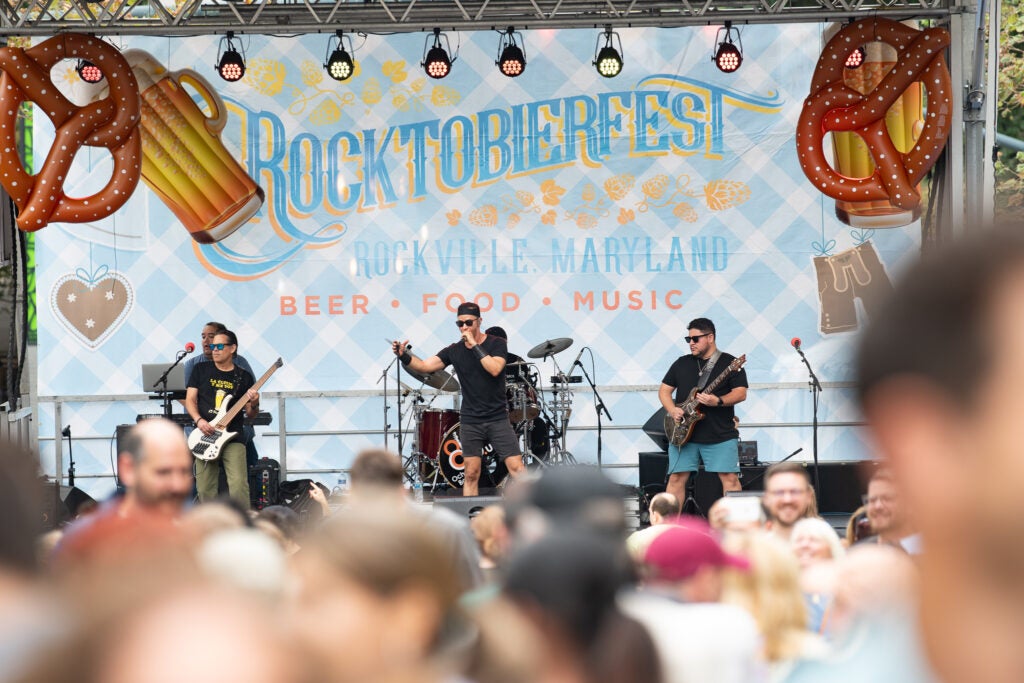 A band performs on stage at Rocktobierfest in Rockville, Maryland, with a crowd watching and a banner advertising beer, food, and music.