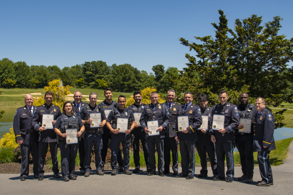 A group of uniformed emergency service personnel stand outdoors in a line, each holding a certificate, with trees and a pond in the background.