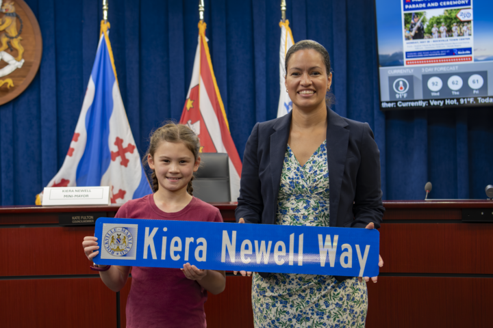 A young girl and a woman stand indoors holding a blue street sign reading 