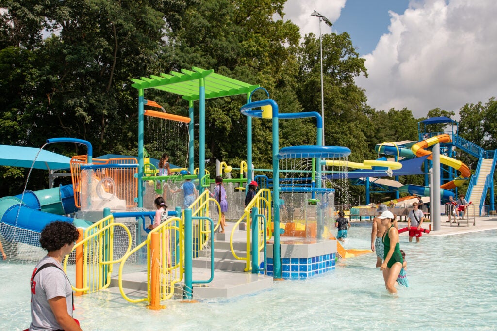Children and adults play at an outdoor water park featuring colorful slides, water sprayers, and climbing structures on a sunny day.