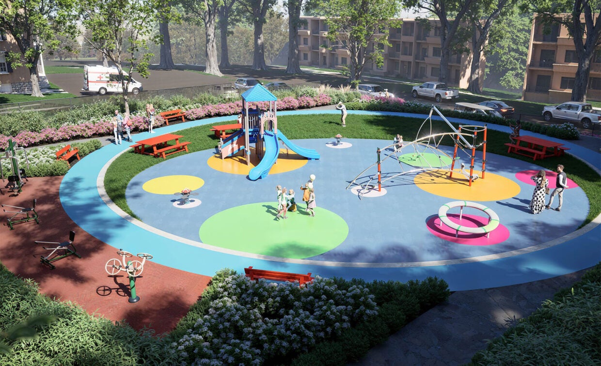 Children and adults gather in a colorful outdoor playground with slides, climbing structures, benches, and greenery, surrounded by trees and parked cars.