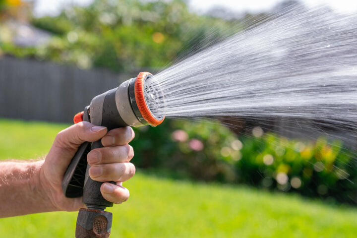 A hand holds a garden hose sprayer, releasing a strong stream of water onto a grassy lawn with blurred plants in the background.