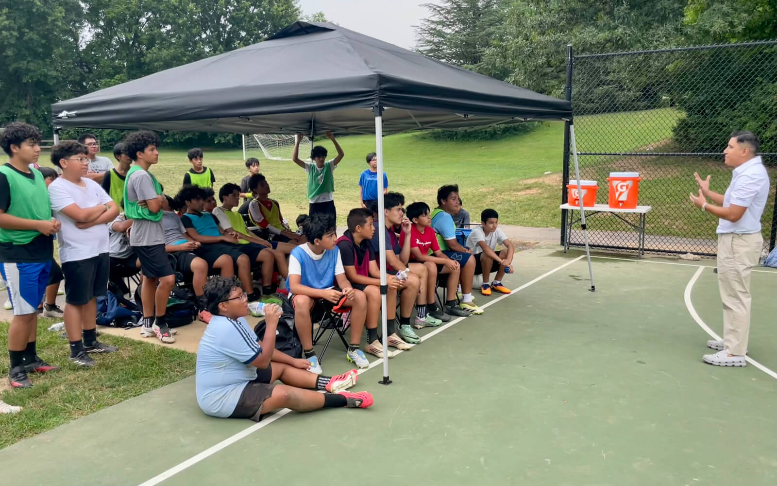 An instructor speaks to a group of young men seated under a canopy on an outdoor sports court, with trees and a fence in the background.