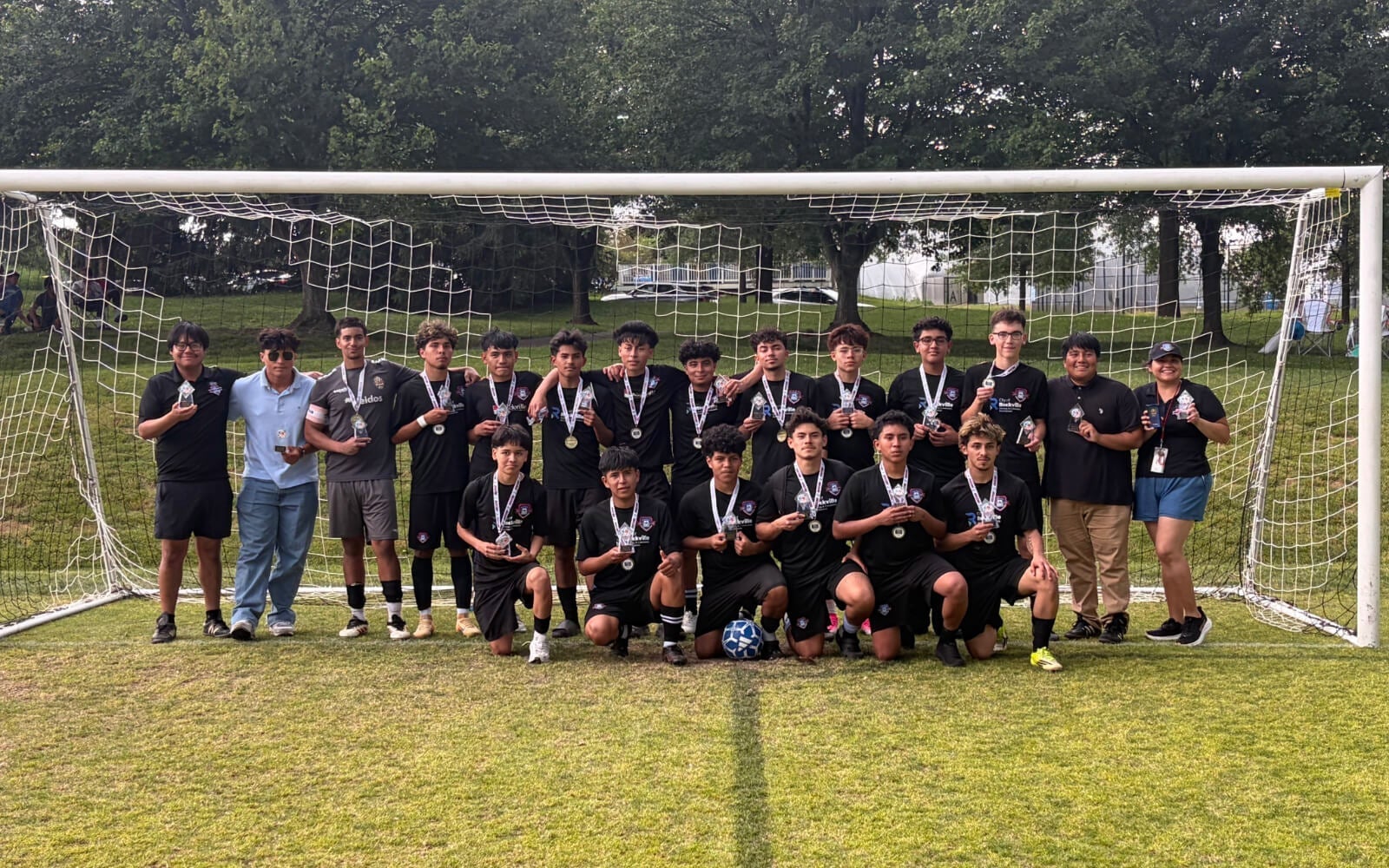 A soccer team poses together in front of a goal on a grassy field, with autumn trees and sunlight in the background.