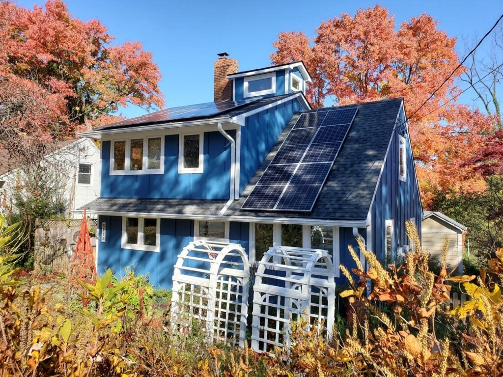 Blue house with solar panels on the roof, surrounded by autumn trees and two white garden arches in the front yard.
