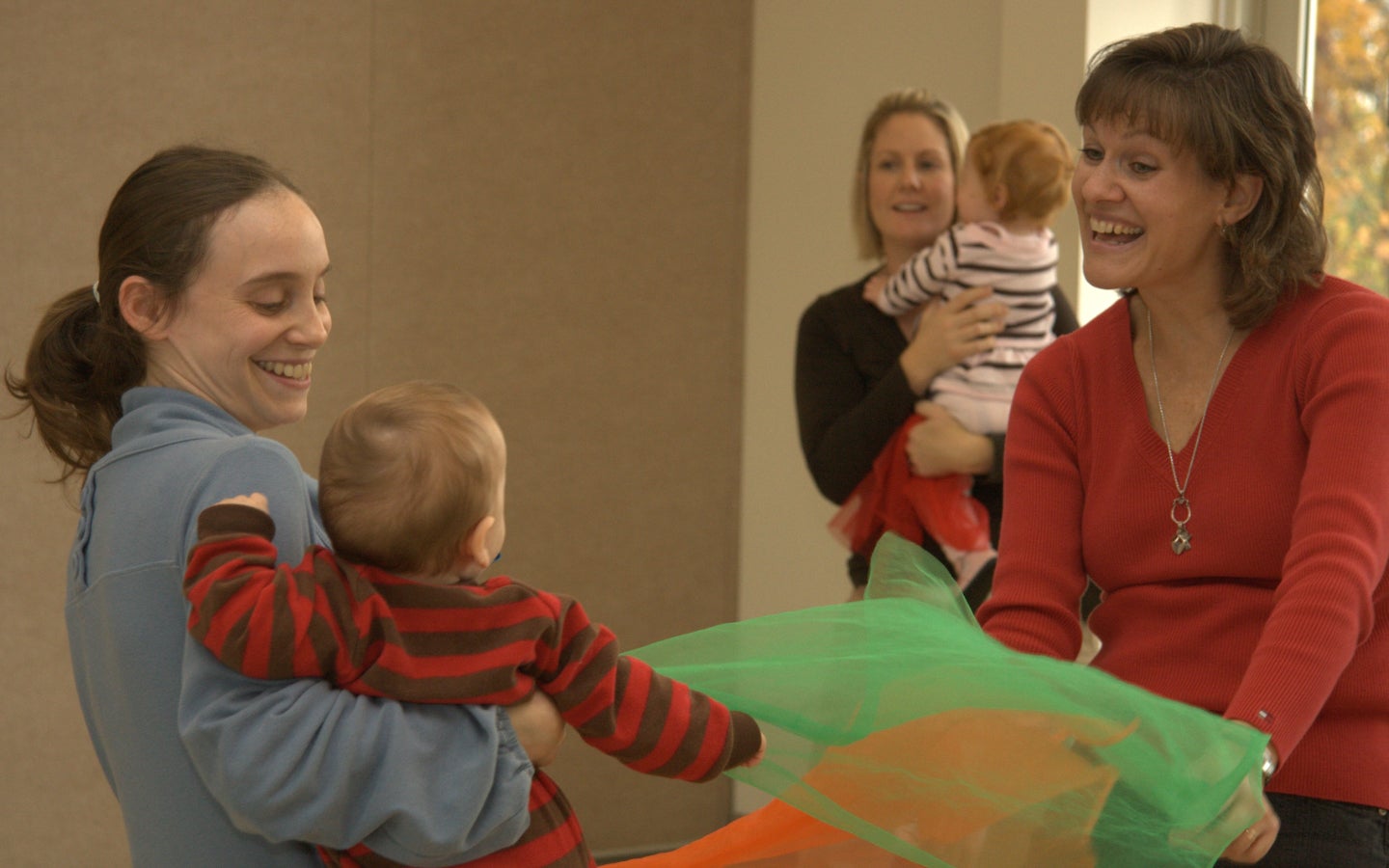 Two women each hold a baby while playing with colorful scarves indoors. Another woman and baby are visible in the background.