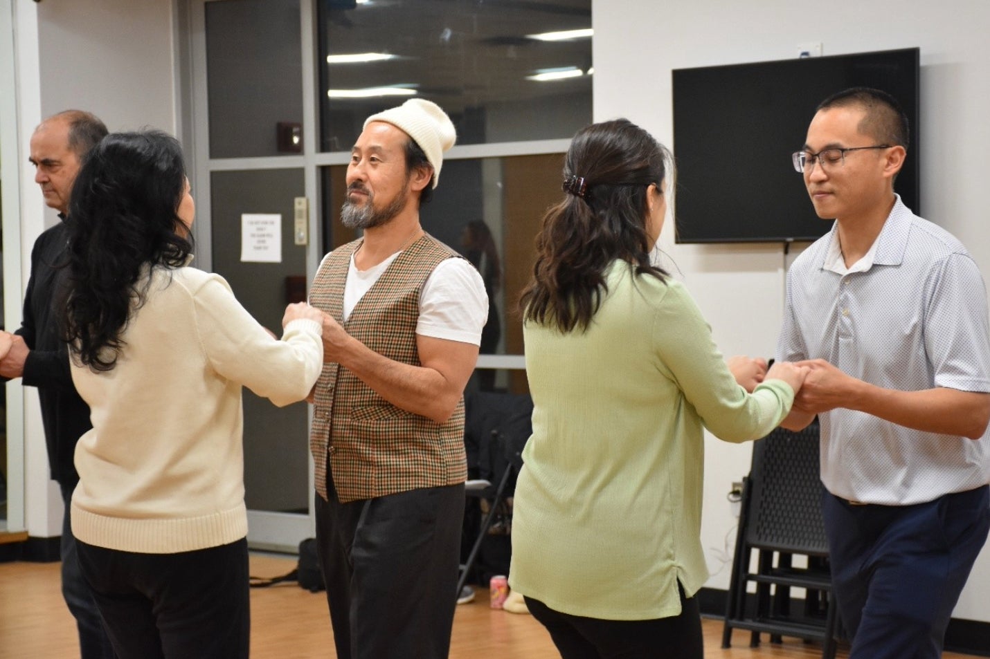 Four people pair up and hold hands while practicing a dance indoors; two other individuals are visible in the background.