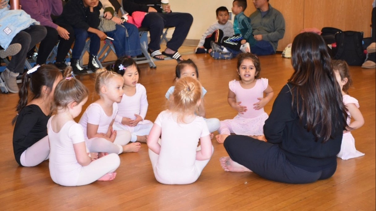 A group of young children in ballet outfits sit in a circle on a wooden floor with their instructor, while adults watch from chairs in the background.
