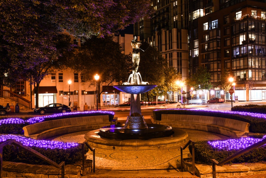 A bronze fountain statue stands in a circular plaza at night, surrounded by purple-lit landscaping and modern buildings in the background.