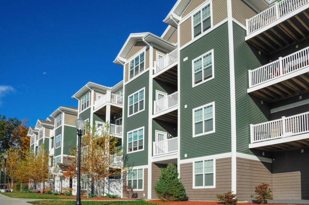 Modern four-story apartment building with green siding, white balconies, and landscaped grounds under a clear blue sky.