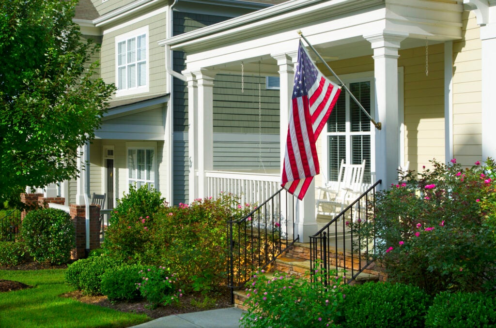 A neatly landscaped suburban house with an American flag hanging from the front porch, featuring bushes, flowers, and a white rocking chair.