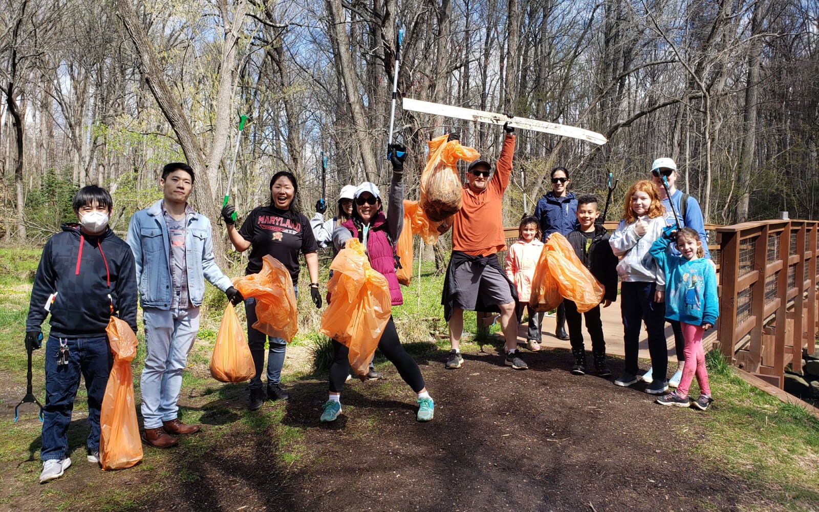 A group of people holding orange trash bags pose together outdoors in a wooded park, participating in a cleanup activity near a wooden bridge.