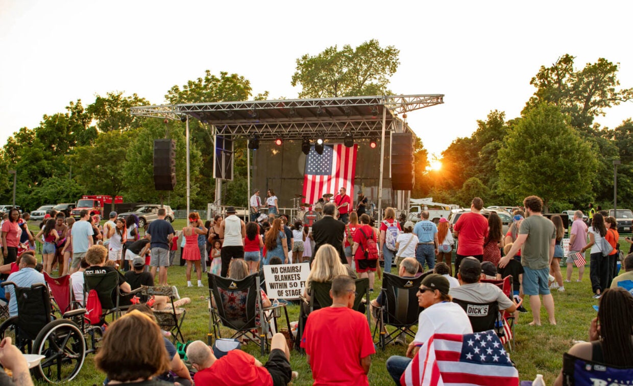 People gather on a lawn facing a stage with a large American flag for an outdoor event at sunset. Some attendees sit in chairs while others stand or walk.