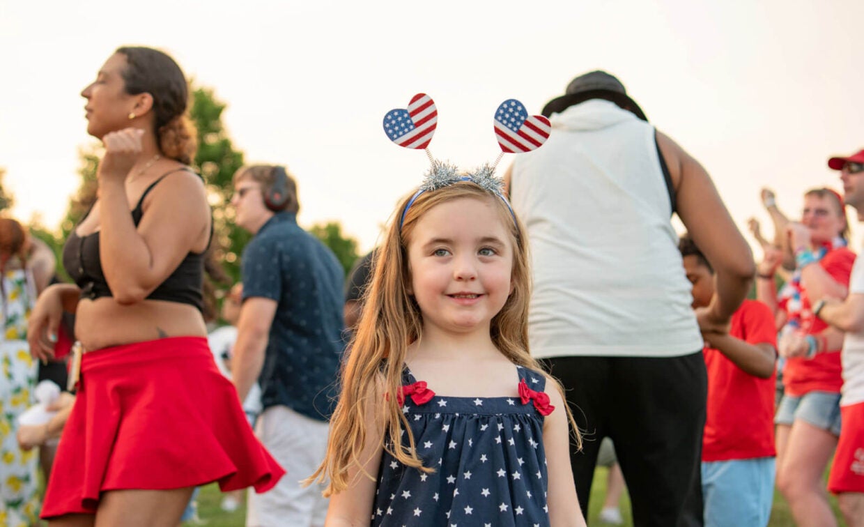 A young girl wearing a star-patterned dress and heart-shaped American flag headband stands outside among a group of people.