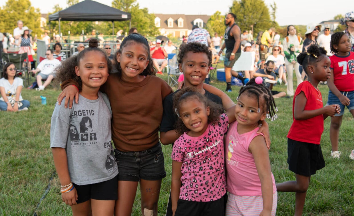 Five children pose and smile for a photo at an outdoor event, with a crowd and tents visible in the background on a sunny day.