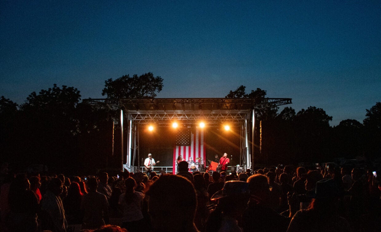 A band performs on an outdoor stage with a large American flag backdrop at dusk, as a crowd watches.