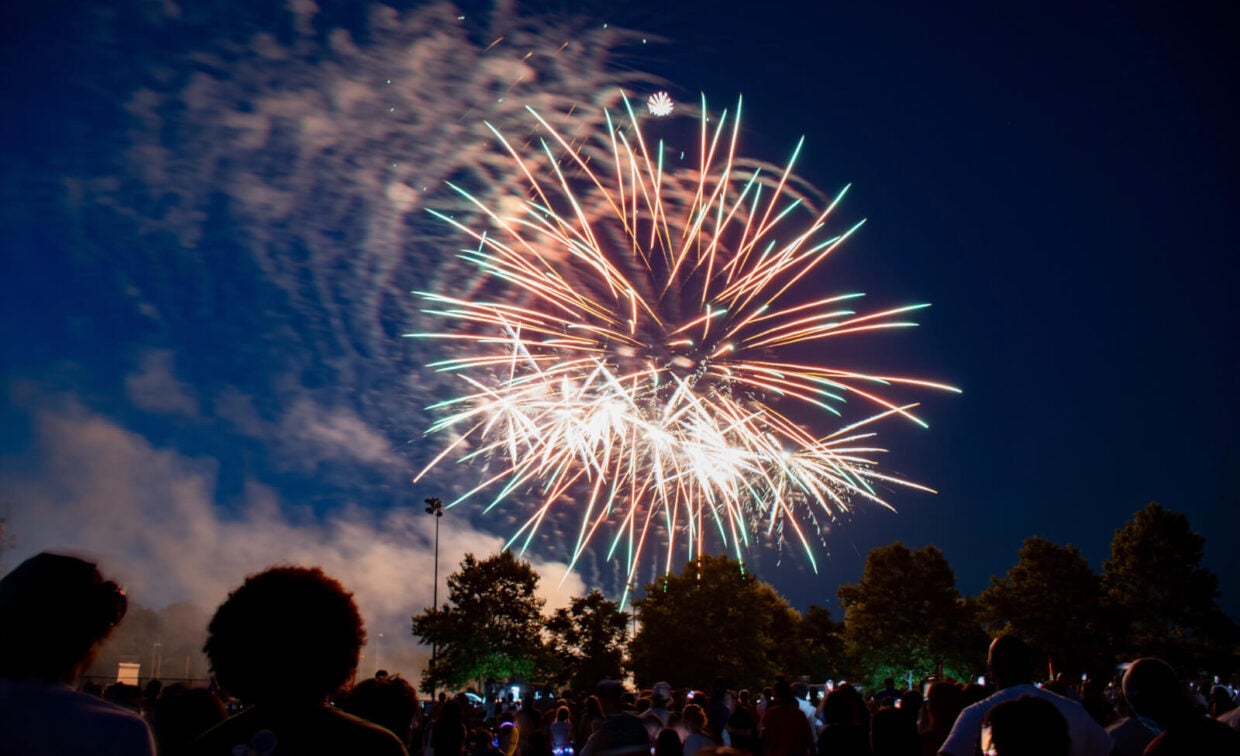A crowd watches colorful fireworks light up the night sky above trees during Independence Day.