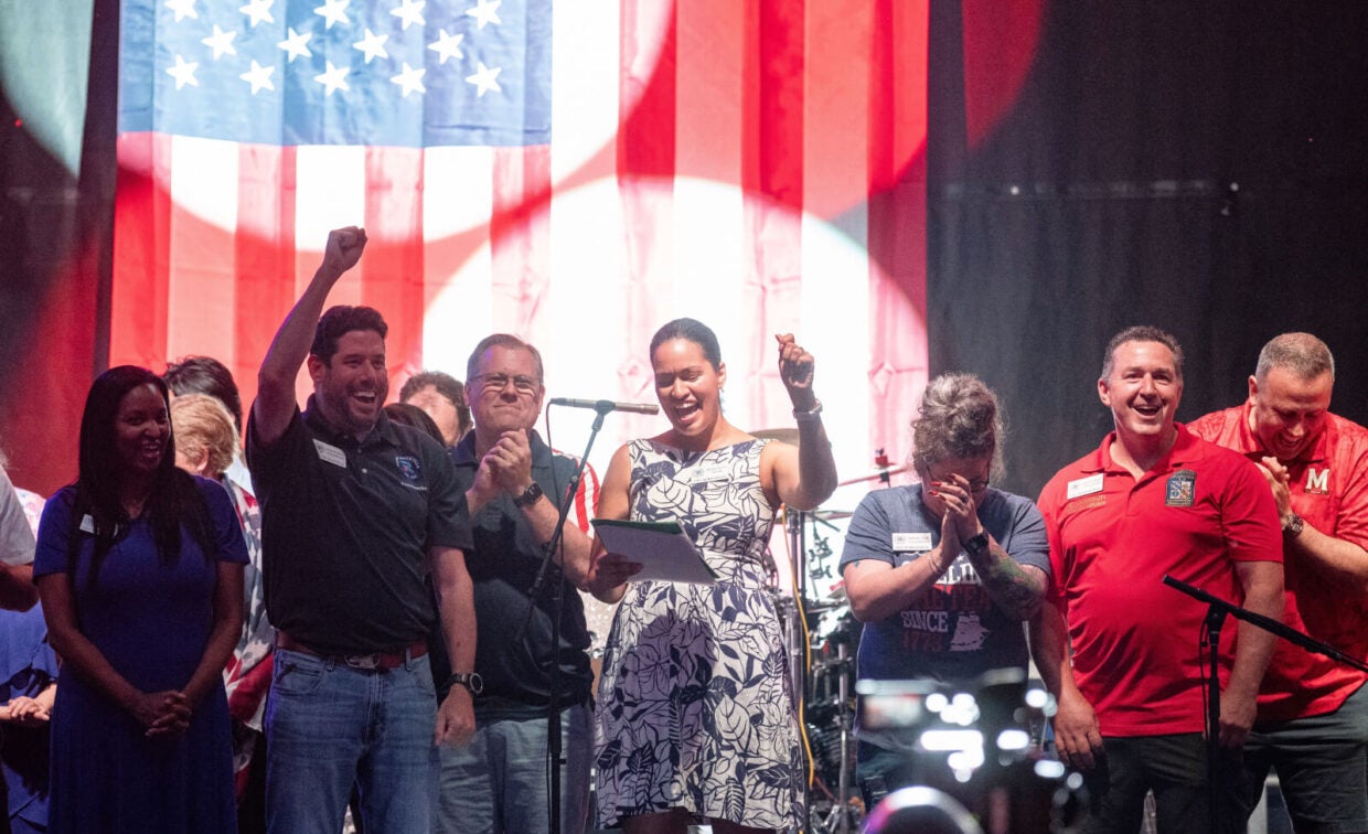 A group of people stand on stage in front of a large American flag; one woman speaks at a microphone while others cheer and smile.