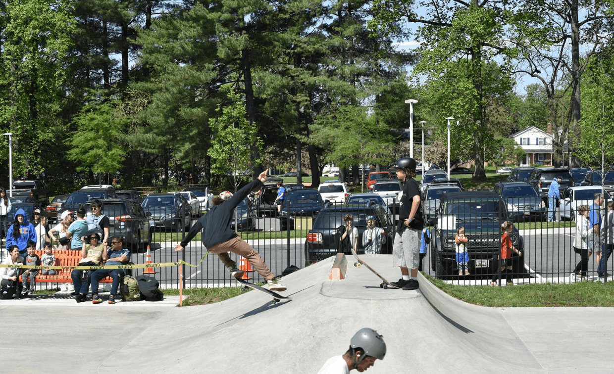 A skateboarder performs a trick on a ramp at a skatepark while others watch and wait their turn. Cars and trees are visible in the background.