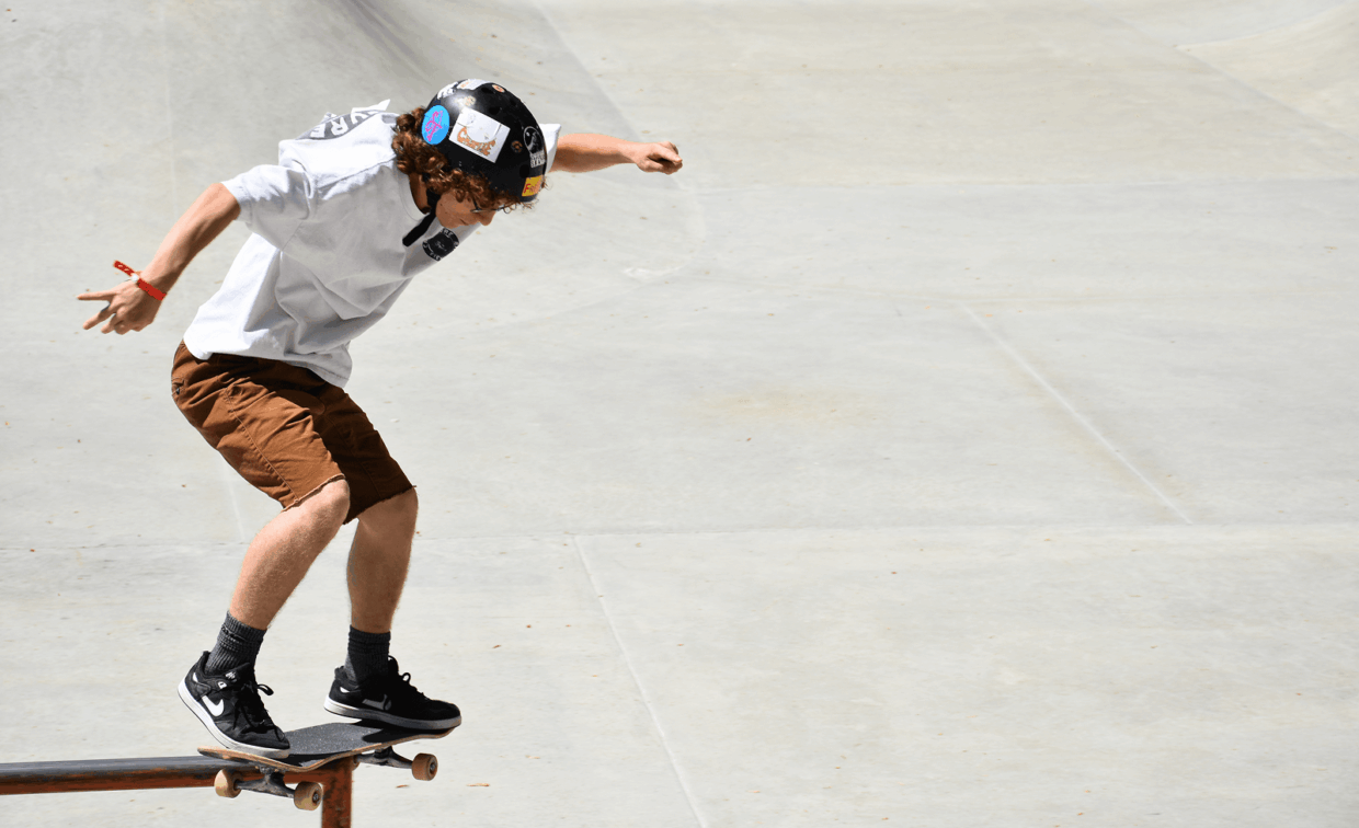 A skateboarder wearing a helmet and shorts balances on a rail at a concrete skatepark during the day.