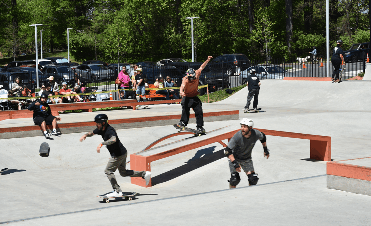 Several skateboarders perform tricks and ride in a busy outdoor skatepark while spectators watch from the sidelines; trees and parked cars are visible in the background.