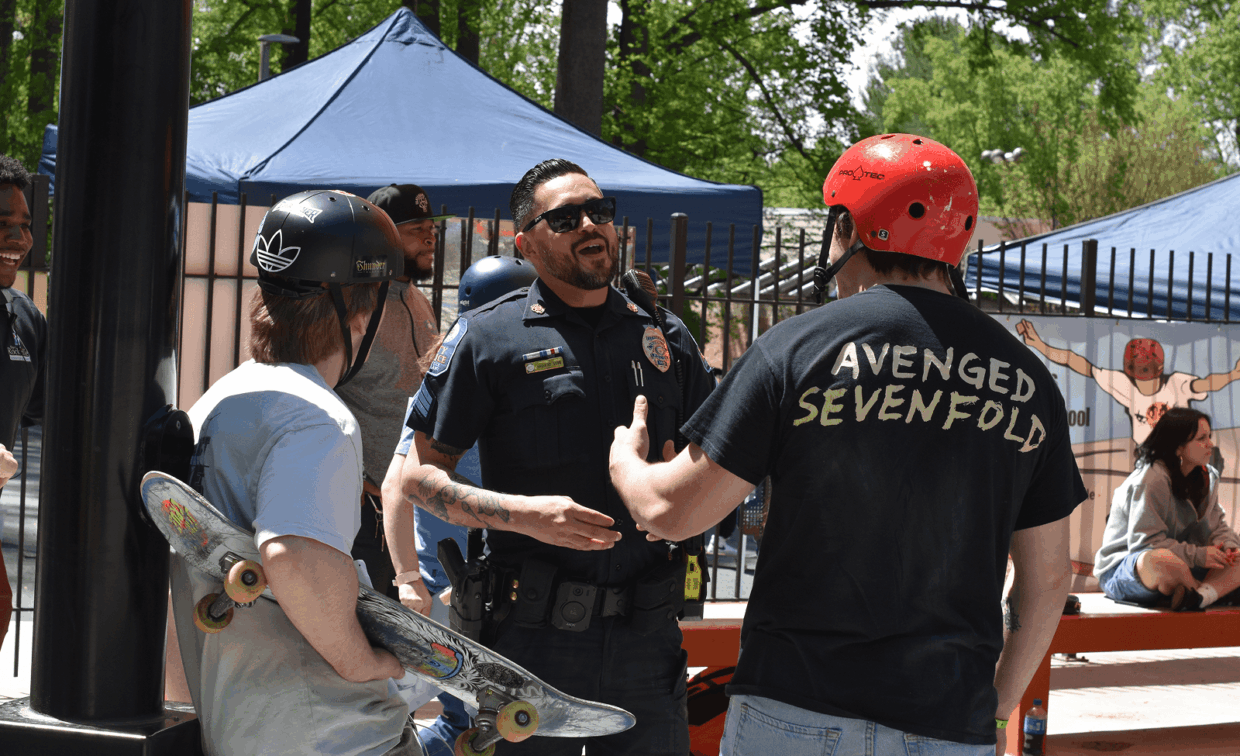 A police officer talks with two skateboarders, one holding a skateboard, at an outdoor event with tents and people in the background.