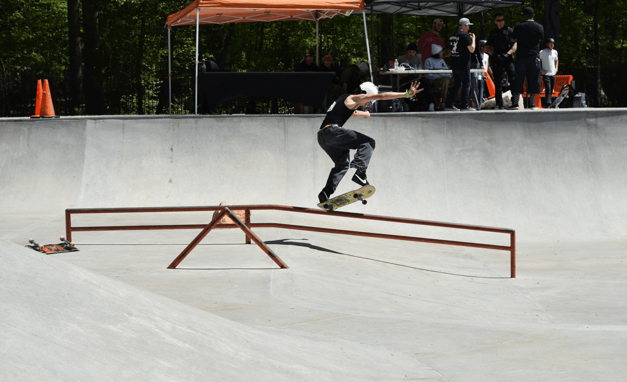 A skateboarder in black clothing rides a rail at a skatepark, with onlookers and judges watching from behind an orange tent.